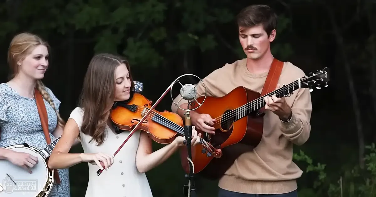 A musical performance outdoors with three young adults, each playing an instrument: a banjo strap, violin and bow, acoustic guitar; lush greenery in background suggests spring or summer with natural daylight.
