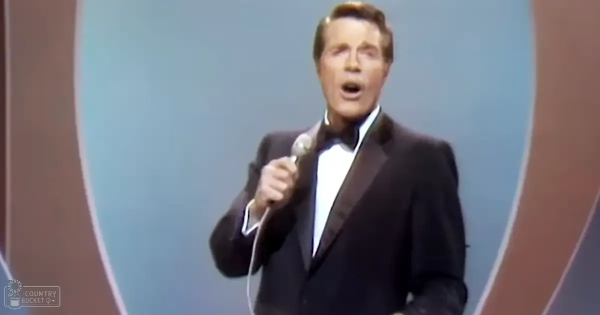 An adult male performer in a black tuxedo, holding a microphone on an indoor stage with blue and brown tones.