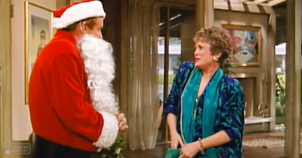 An adult male dressed as Santa Claus interacts with a woman wearing a blue patterned top and wristwatch in an indoor living room setting, likely during winter.