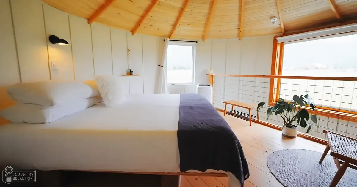 A rustic cabin interior with white bedding, wooden furniture, and a view of an open green landscape during daylight.