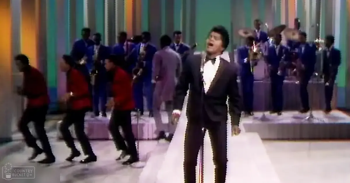 A male adult performer in a dark suit jacket and white shirt singing on stage with young adult male dancers wearing red jackets and synchronized musicians in blue suits holding brass instruments.