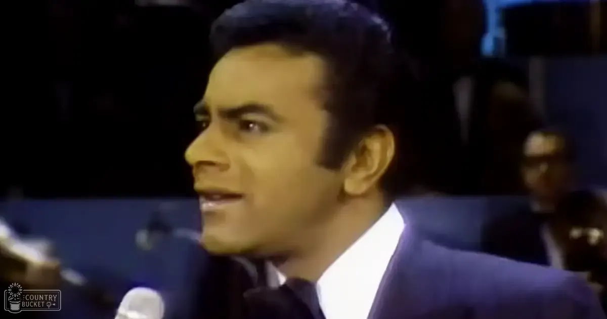 A young adult male wearing a dark blue suit, white shirt, and tie stands indoors on what appears to be a stage or studio with blurred figures in formal attire in the background.