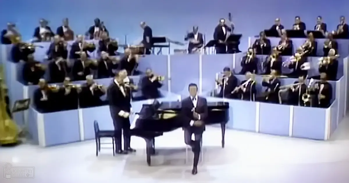 A musical performance with a conductor seated at the piano, surrounded by an orchestra of adults dressed uniformly in dark suits and white shirts, set against a plain light blue backdrop under artificial stage lighting.