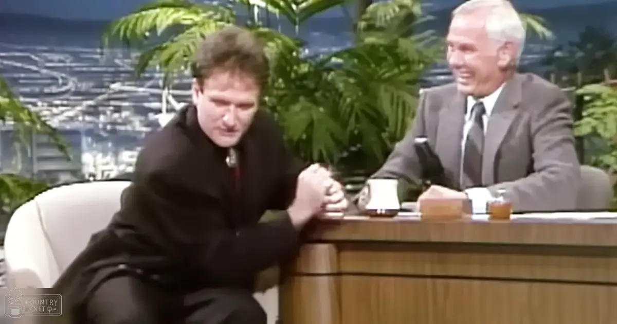 A late-night talk show set with two male hosts seated at a desk against an out-of-focus cityscape backdrop with large green plants.