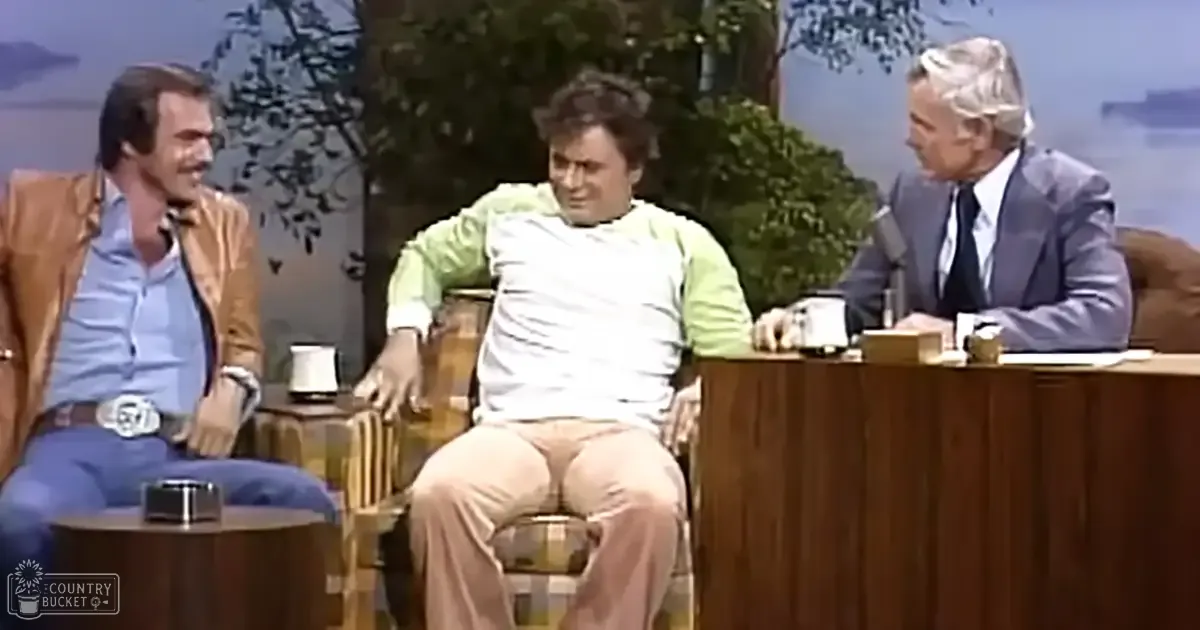 Adult male in a brown jacket seated with two other men, likely part of a talk show set; backdrop features painted trees and water suggesting an outdoor scene.