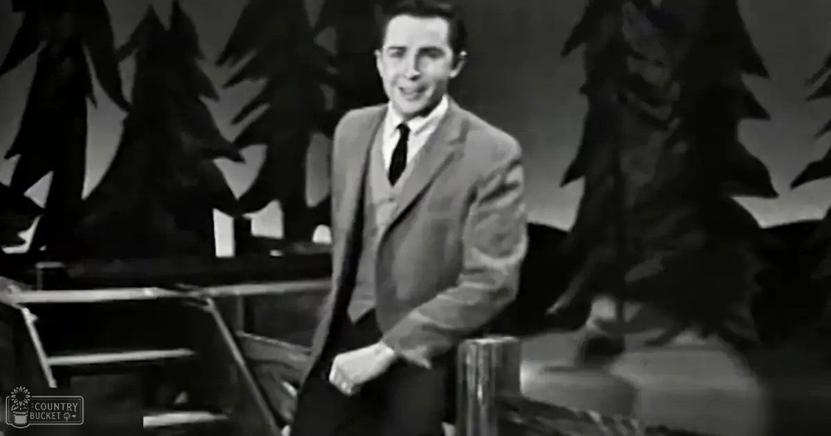 A young adult male wearing a dark suit jacket with a lighter-colored shirt and tie in an indoor setting, possibly a studio or stage.