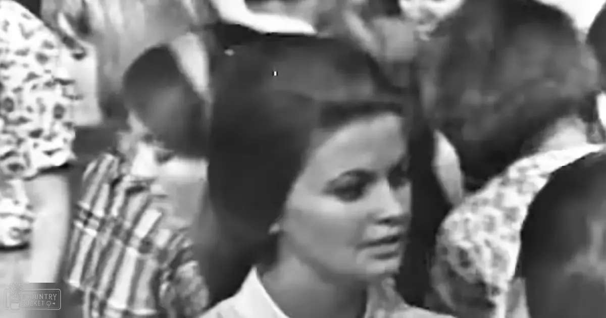 A young adult female with dark hair styled back stands outdoors wearing a white garment with a high neckline and short sleeves; behind her, a child or toddler in muted striped shirt is partially visible. The setting appears to be an outdoor crowd area during daytime.