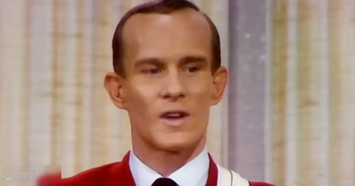 A young adult male wearing a red sweater vest over a white collared shirt and dark tie, standing indoors with light-colored curtains in the background.