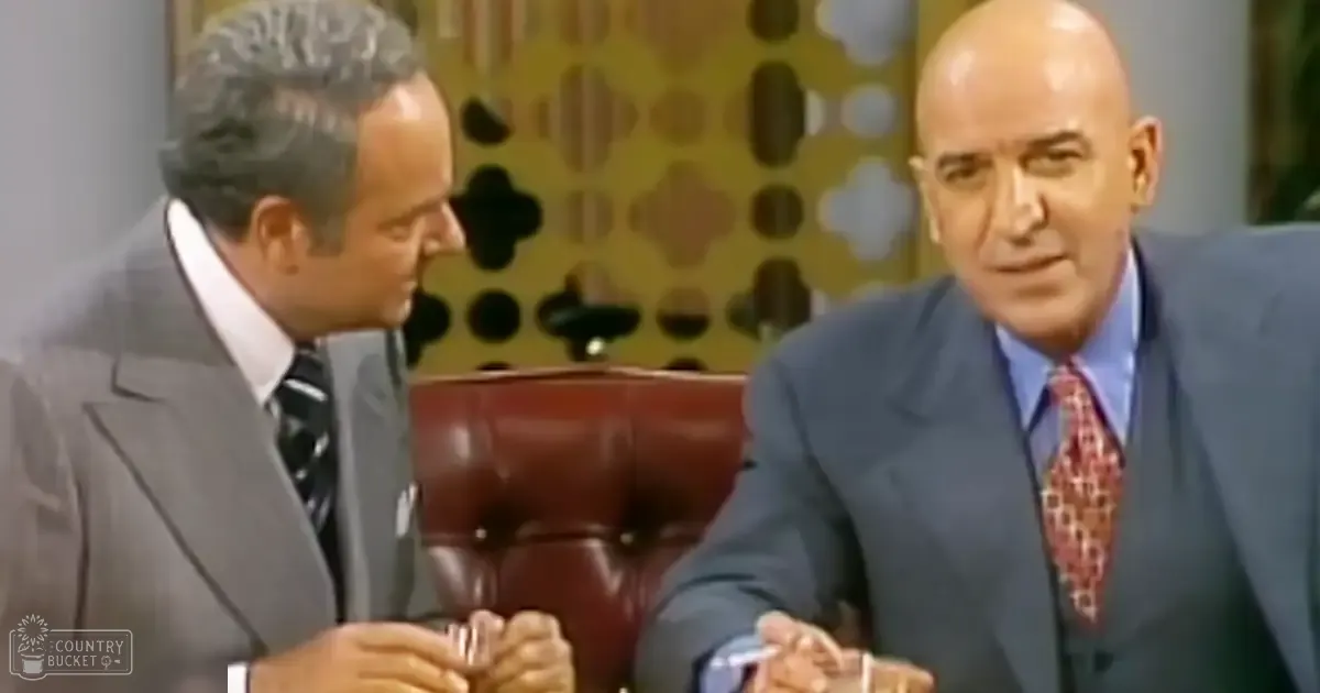 Talk show set with two adult men in formal attire; studio environment, abstract wall pattern, even lighting.