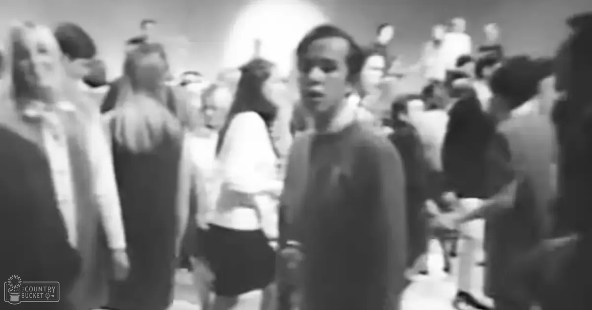A young adult male stands at the center indoors with others around him, wearing a dark-colored garment in an even-lit room.