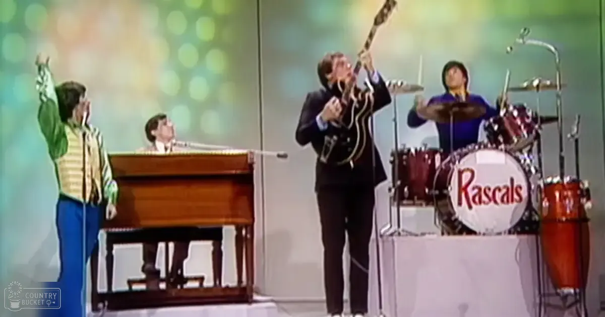 A musical performance with 'The Rascals' in a studio setting; young adult male singer wears green and yellow striped shirt with long sleeves, seated at a keyboard or piano is in light-colored suit from the mid-20th century, another young adult male rock musician holds guitar wearing dark suit typical of late 1960s or early 1970s fashion, adult male drummer plays 'Rascals' branded drum set with blue long-sleeve shirt and dark pants; backdrop is simple light-colored screen with bokeh effects.