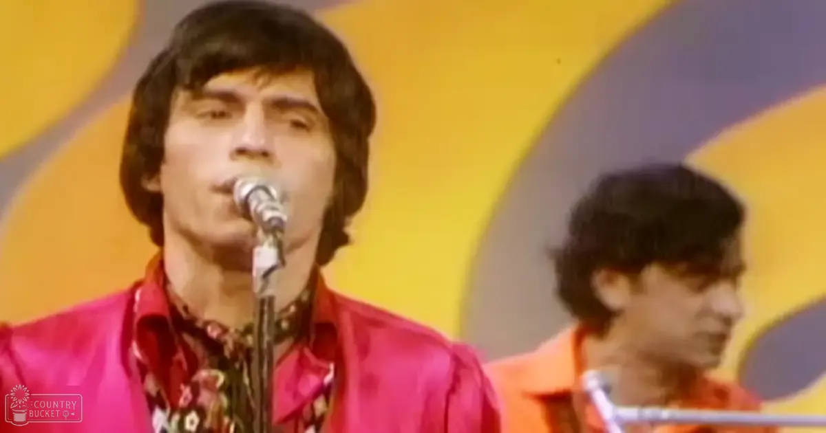 A young adult male singer performing at a music venue, surrounded by bright red and orange shirts with floral patterns against a yellow backdrop.