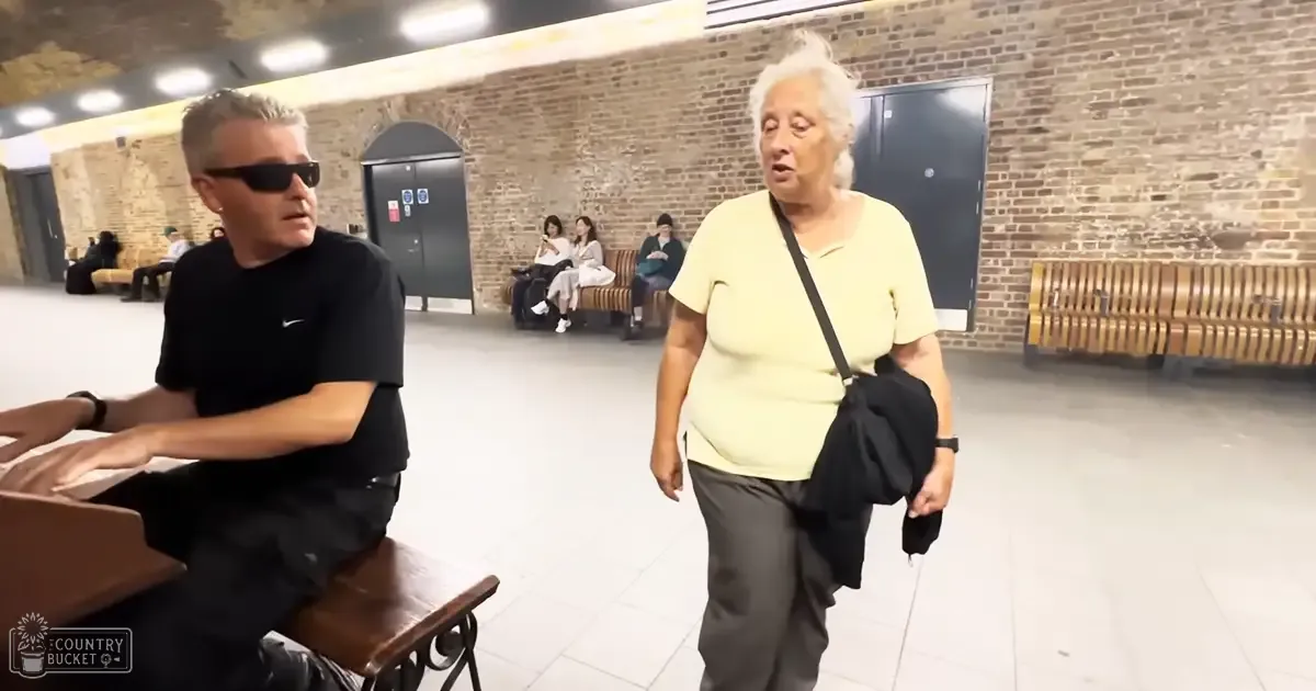 Middle-aged man seated at a piano in an indoor public space with brick walls; older adult woman nearby carrying a black crossbody bag.