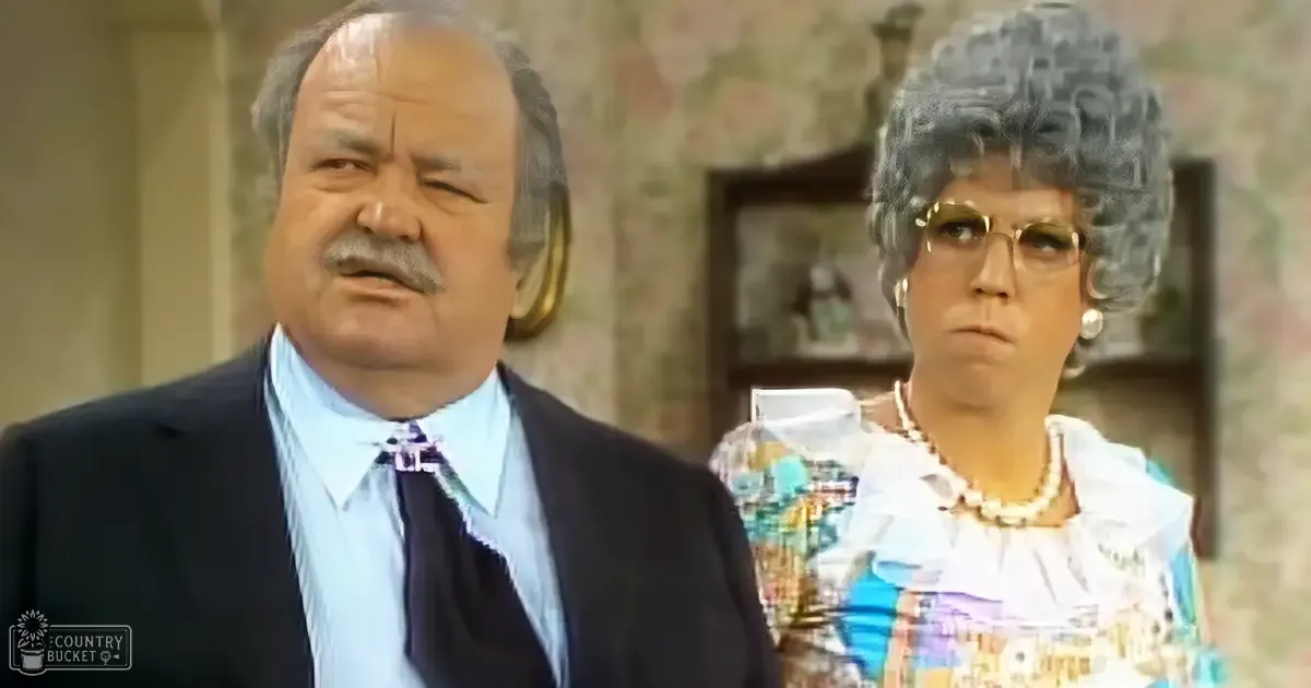A middle-aged man wearing a dark suit jacket over a light blue dress shirt with a dark tie, paired with an older adult woman in a colorful patterned blouse and pearl accessories, standing indoors with vintage decor.