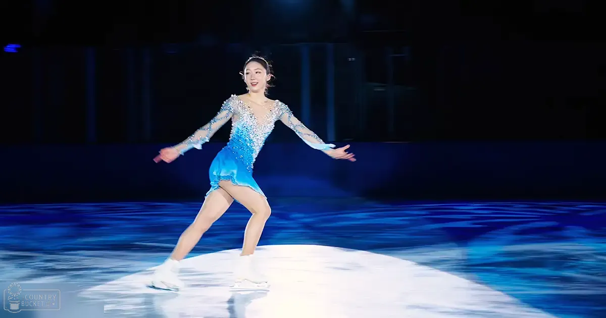 A young adult female figure skater in a blue skating costume with intricate silver embellishments performs on an indoor ice rink under dramatic blue lighting.