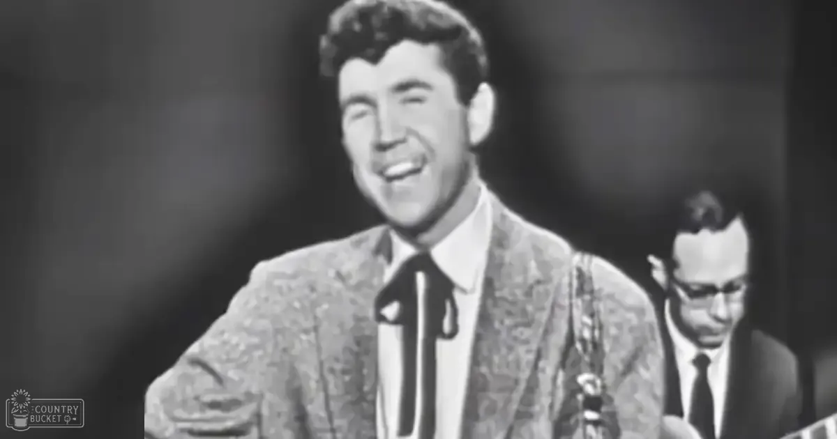 An adult male performer in a patterned suit jacket and white shirt, with a dark-suited background person wearing glasses, set against a black-and-white studio backdrop.