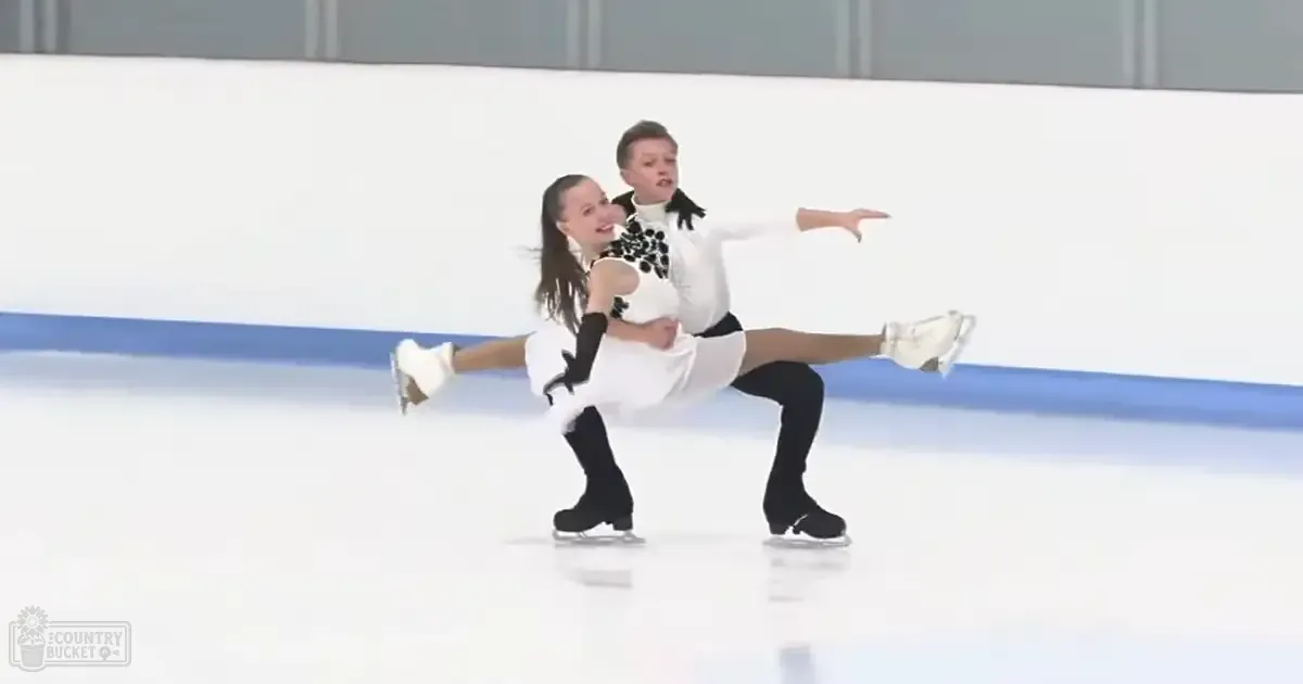 A figure skating duo, a female and male teenager dressed in white skirts with black floral patterns and black tights, performing on an indoor ice rink under artificial lighting.