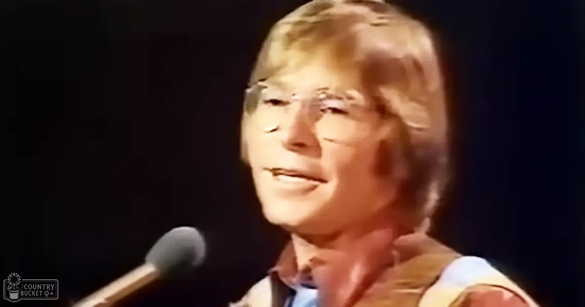 An adult male singer wearing a light-colored, short-sleeved collared shirt with rolled-up sleeves and a blue tie in front of a microphone on stage.
