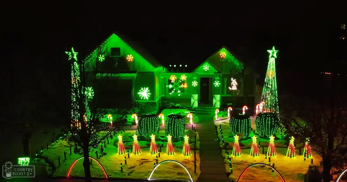 A house adorned with green and white Christmas lights, including snowflake patterns on the roof and walls, surrounded by small illuminated trees and candy canes. The scene is set in an urban residential area during winter.