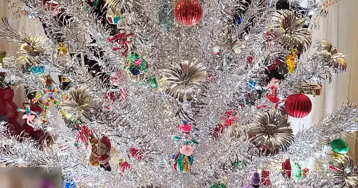 A decorated Christmas tree indoors, surrounded by various holiday-themed ornaments under indoor lighting during daytime or early evening.