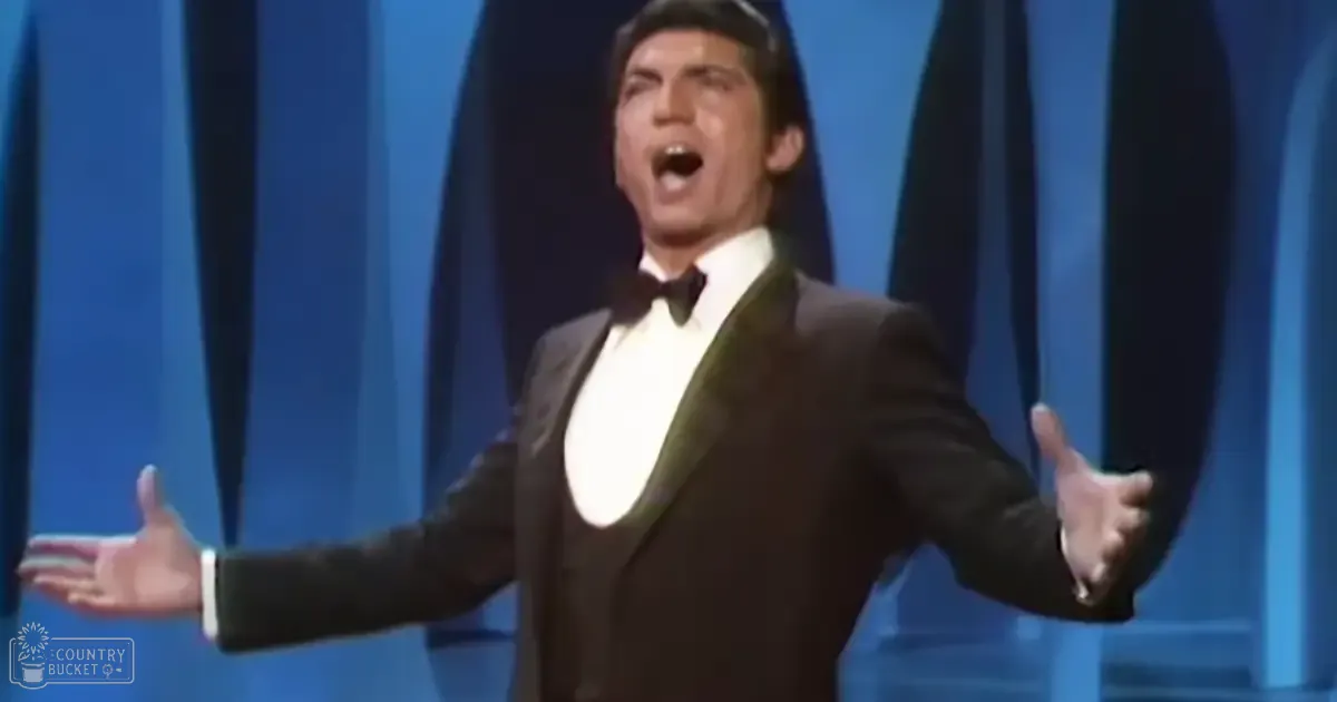 An adult male performer in a black mid-20th-century tuxedo with a white shirt and black bow tie, set against a blue curtain backdrop suggesting a theatrical stage.
