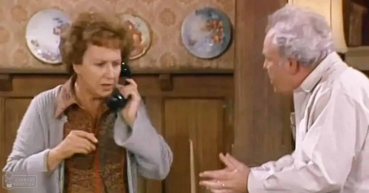 A middle-aged female wearing a light gray cardigan over a brown patterned blouse, holding a telephone to her ear in a home setting with wooden paneling and decorative plates on the wall. An older adult male in a white shirt is also present.