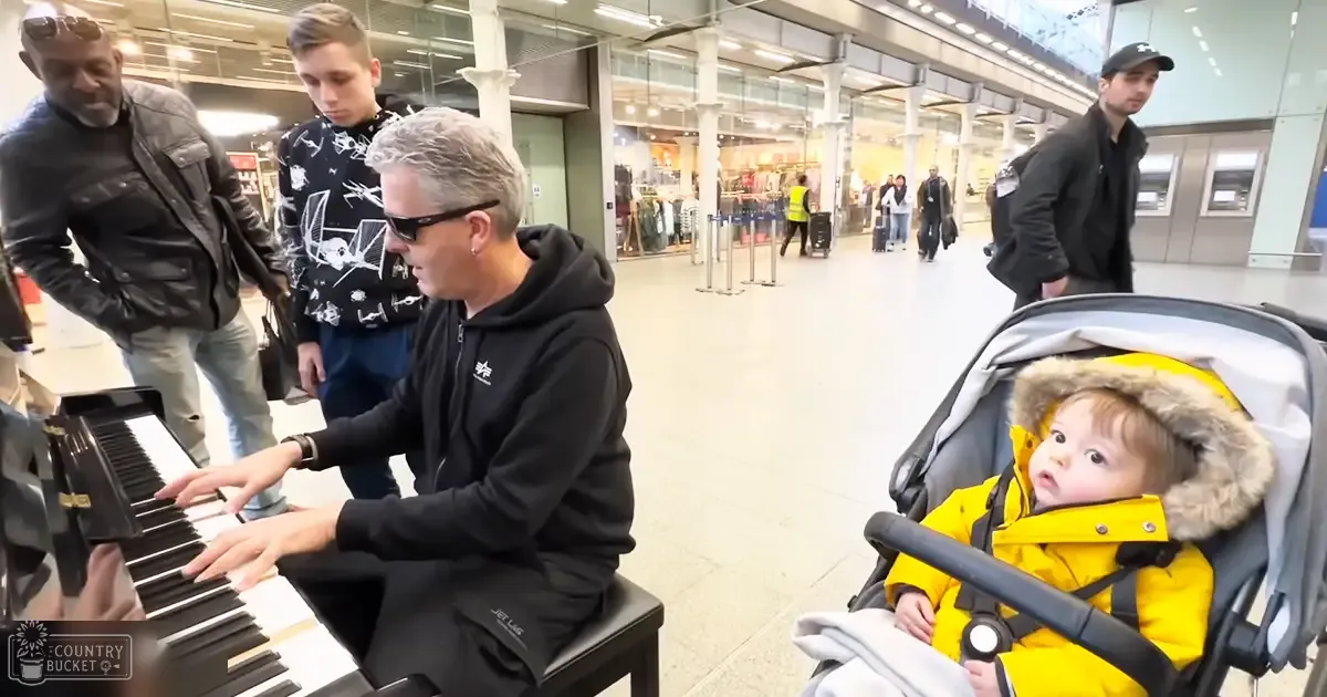 Middle-aged male playing a standard upright piano in an indoor public space resembling an airport terminal, with various individuals and a stroller present around him.