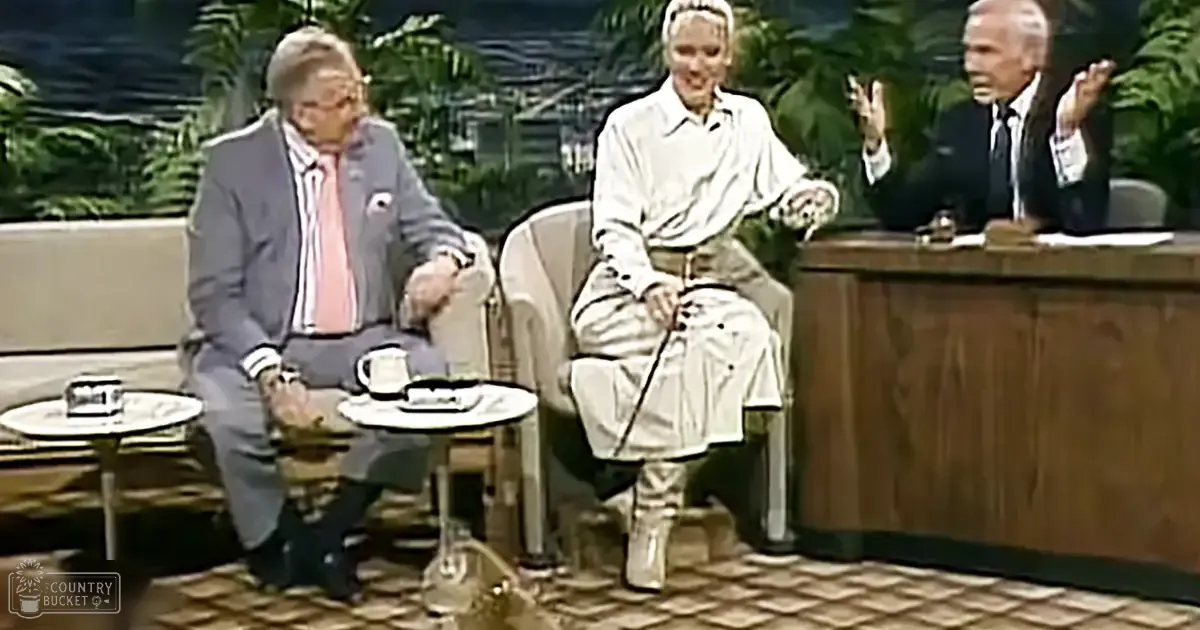 Three individuals in a talk show setting with green foliage backdrop; adult male wearing gray blazer over white shirt and pink tie, adult female holding cane, older male gesturing.