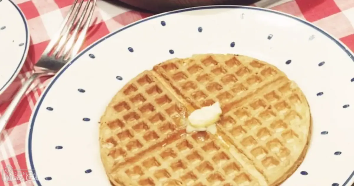 A buttered waffle on a polka-dotted plate atop a red and white checkered tablecloth in an indoor dining area with natural light, likely spring or summer.