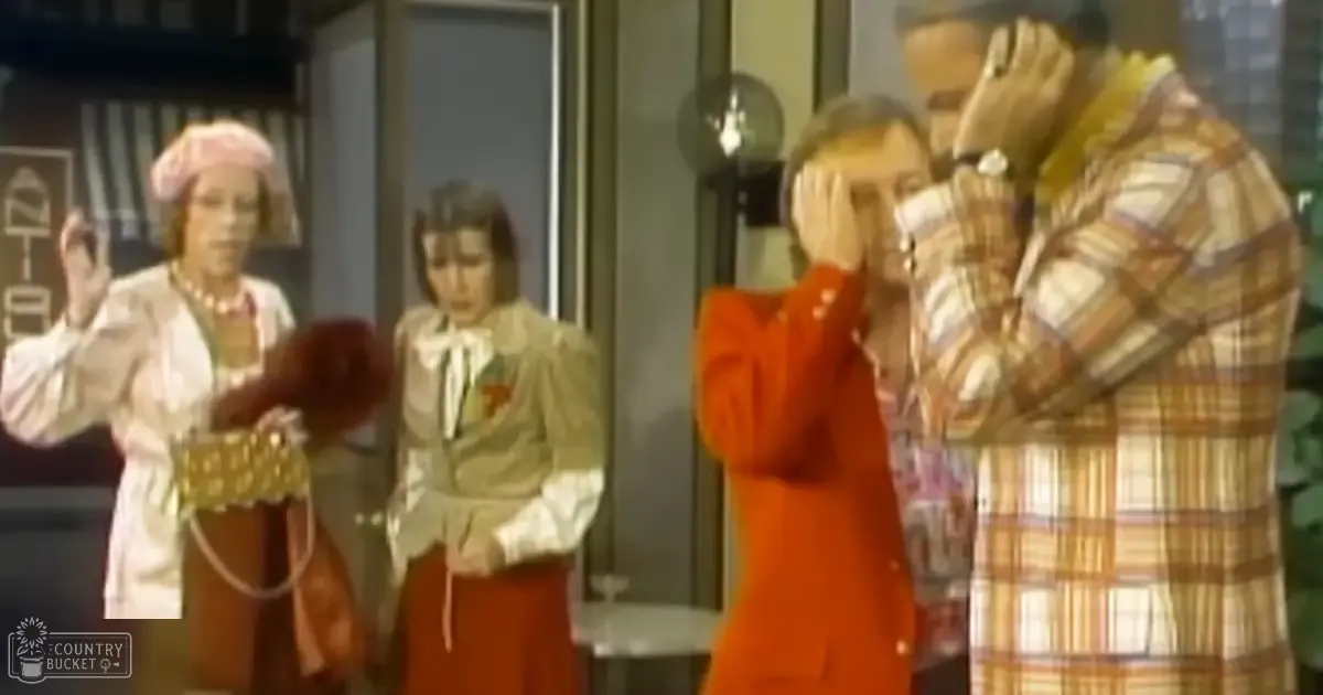 Adult female in a white blouse with gold trim skirt and pink headscarf interacts with another adult female in an orange skirt and white bow tie, alongside an adult male in a plaid shirt indoors with modern decor.