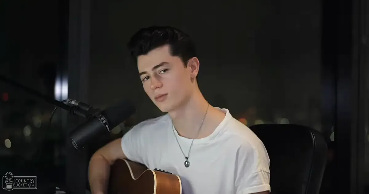Young adult male wearing white t-shirt with relaxed fit and holding acoustic guitar in indoor studio setting, suggesting music recording or performance.