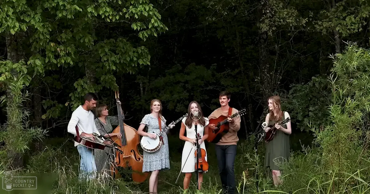 A musical group of six adults performing outdoors, surrounded by trees and greenery under soft daylight.