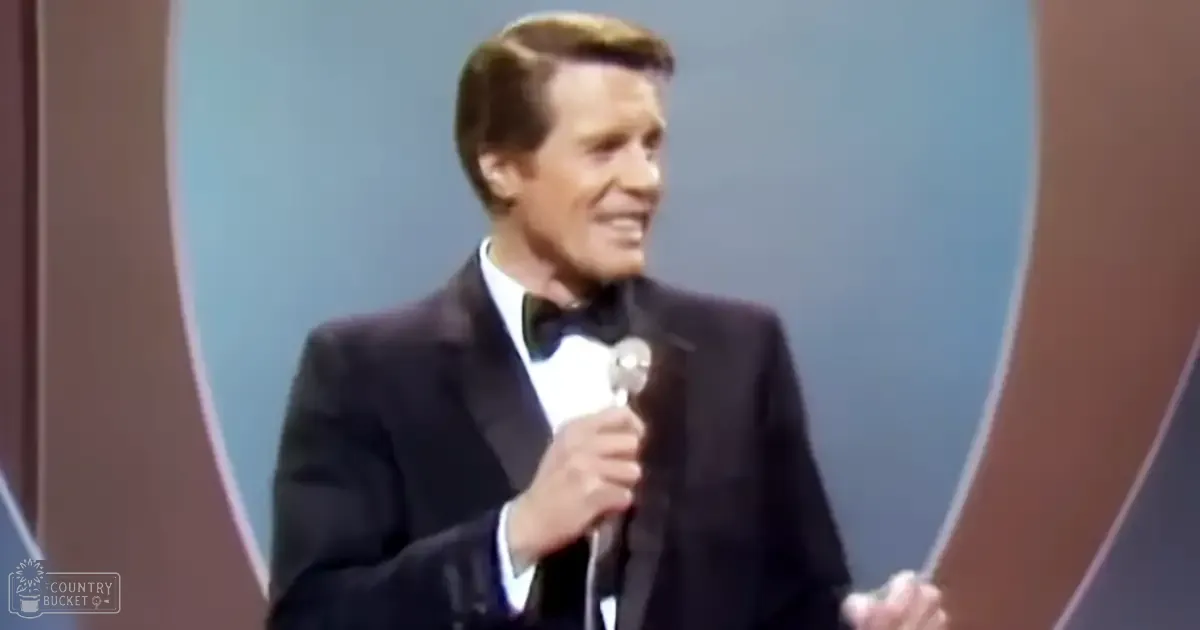 An adult male performer wearing a formal black tuxedo suit with a bow tie, holding a microphone in an indoor stage or studio setting with circular backdrop and muted colors.