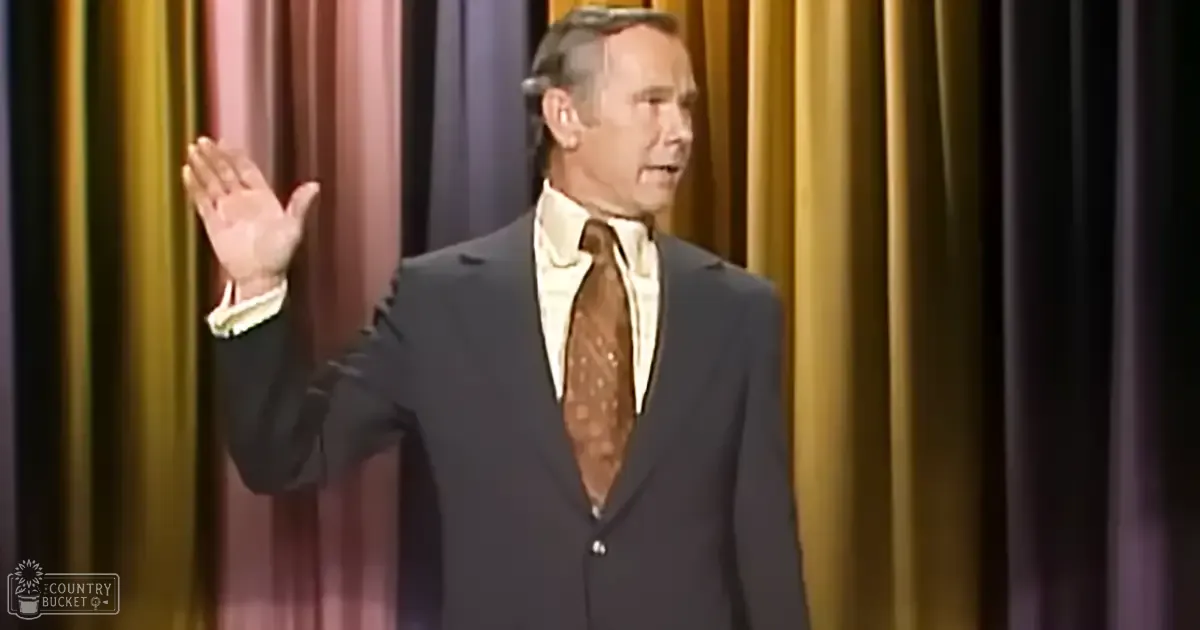 A middle-aged man in a dark suit and light-colored shirt stands on what appears to be a stage or performance area, set against a curtain with vertical stripes in gold and purple. The image suggests an indoor venue with controlled lighting conditions.