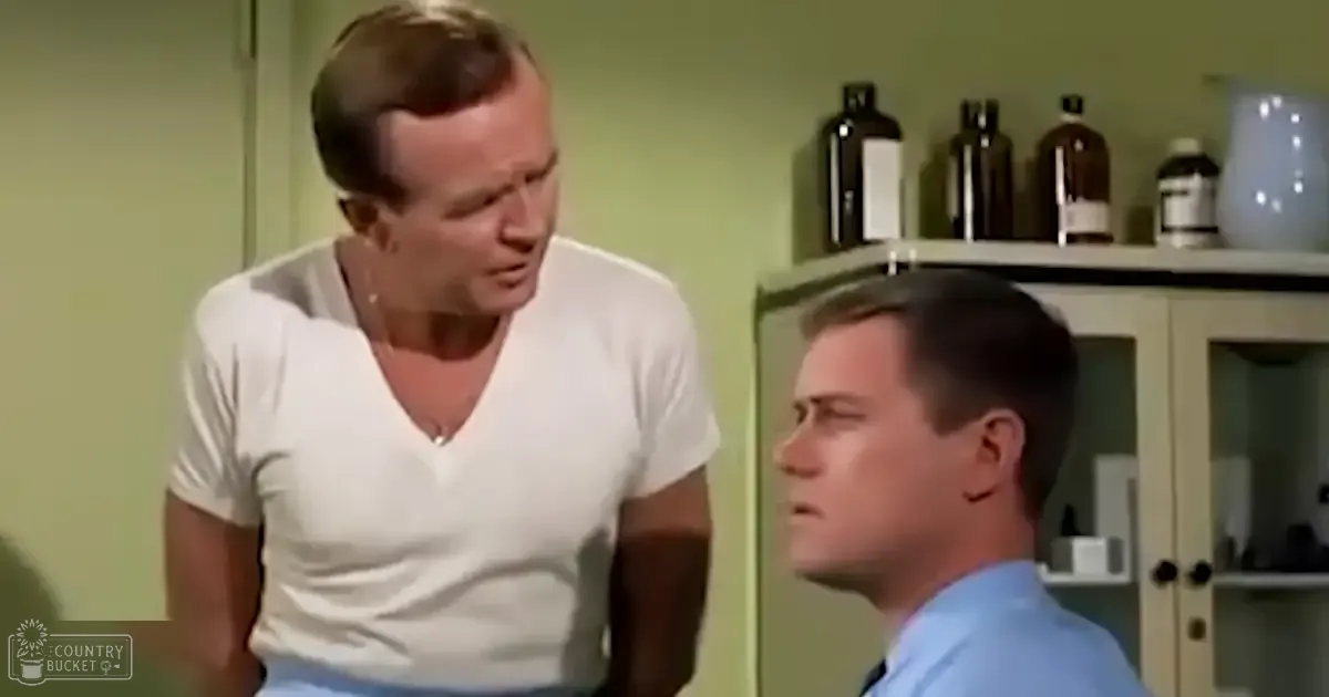 A middle-aged man wearing a white V-neck t-shirt stands next to a young adult male in a light blue button-up shirt, both in an indoor room resembling a laboratory or medical office with glass-cabinet shelves holding dark bottles.