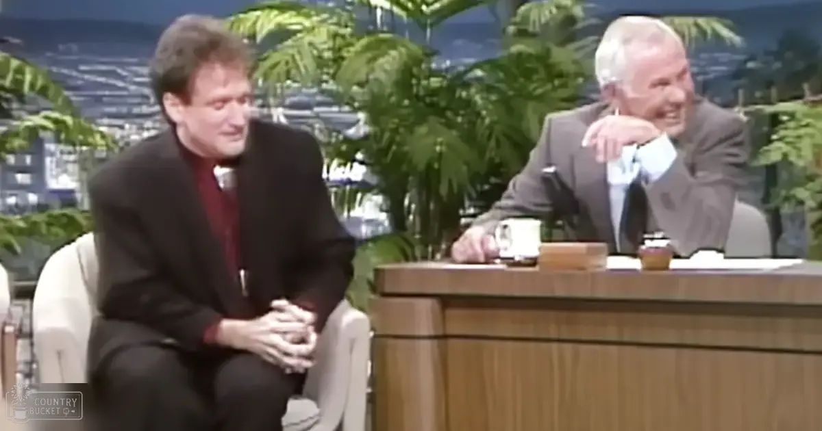 A talk show host and guest seated at a desk in front of an outdoor backdrop with greenery, during what appears to be spring or summer.