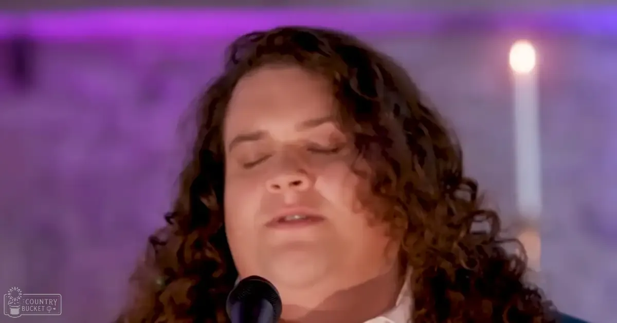 A young adult female singer wearing a loose, white collared shirt with long sleeves, performing into a microphone under purple stage lighting during an indoor event.