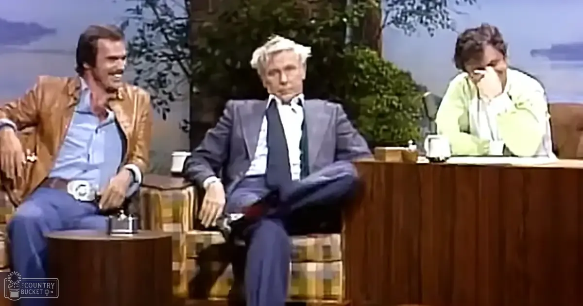 A scenic outdoor setting viewed through windows, with a talk show set inside with three formally dressed men seated at wooden tables.