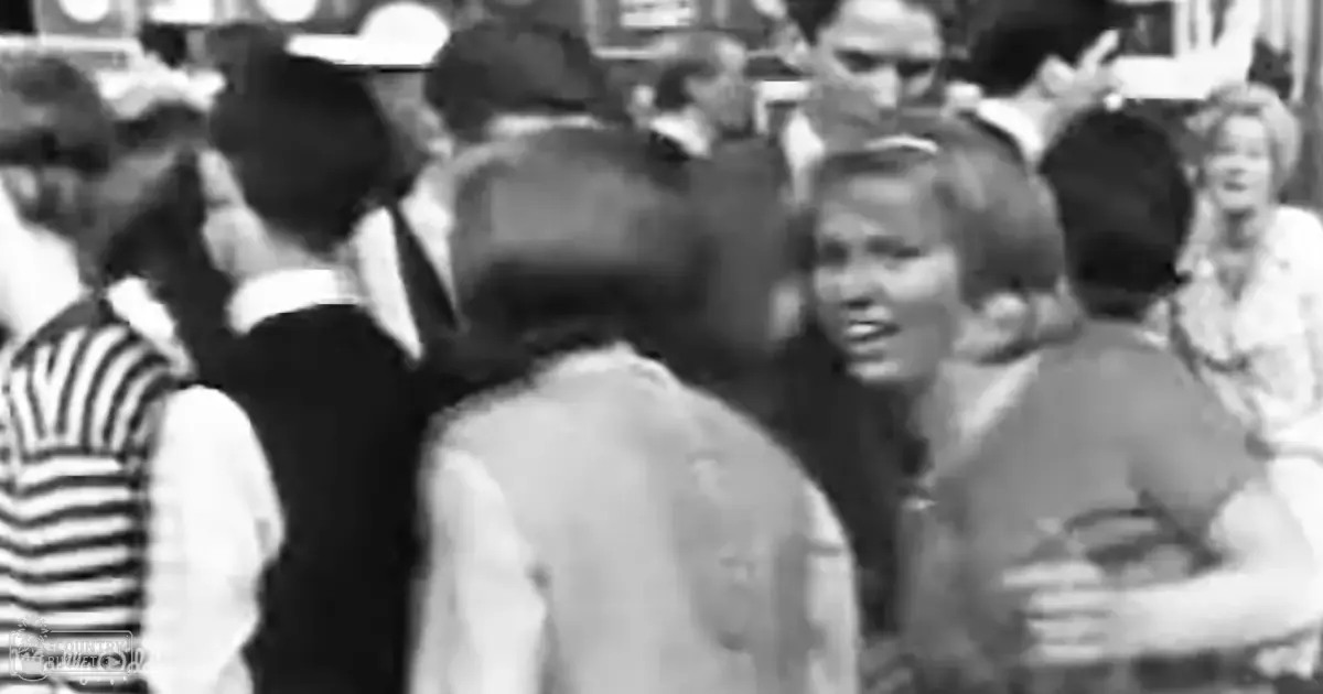 Adult female wearing a light-colored blouse or jacket at an outdoor social event with a crowd in the background; muted daytime lighting.