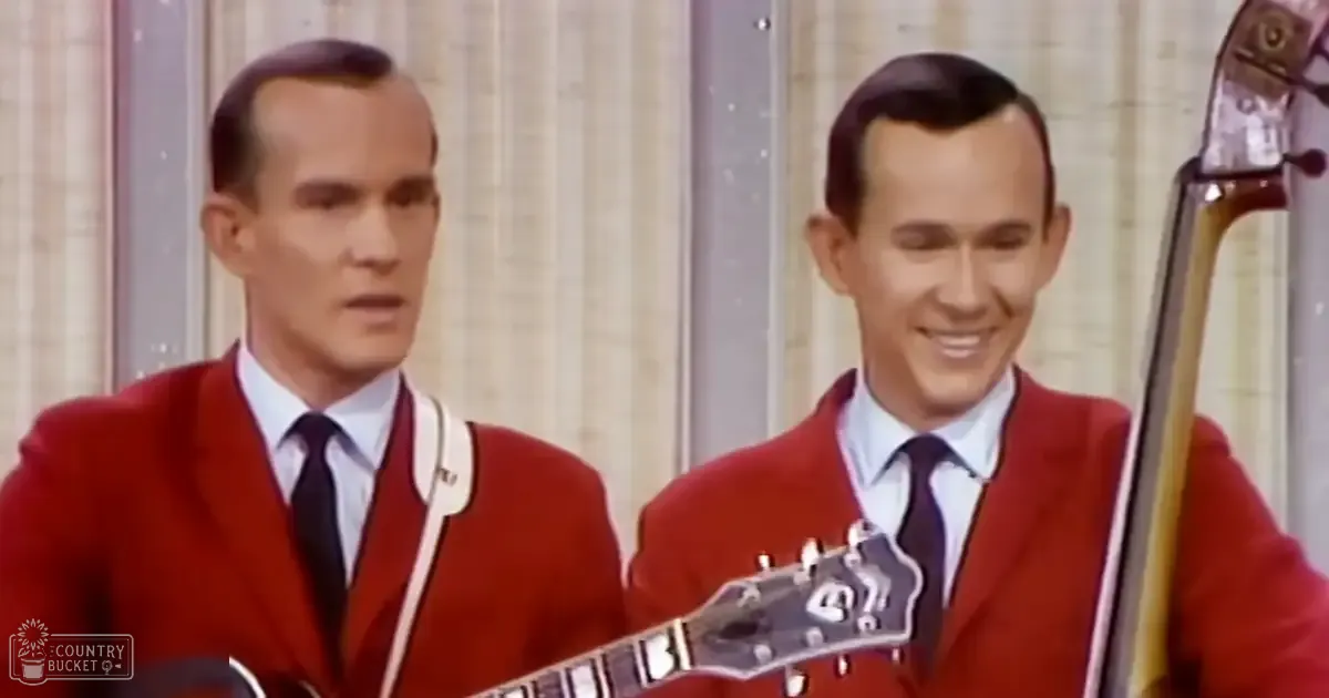 A mid-20th century musical performance scene with two adult male singers in red blazers, white collared shirts, and dark ties, holding stringed instruments against a backdrop of vertical blinds or curtains.