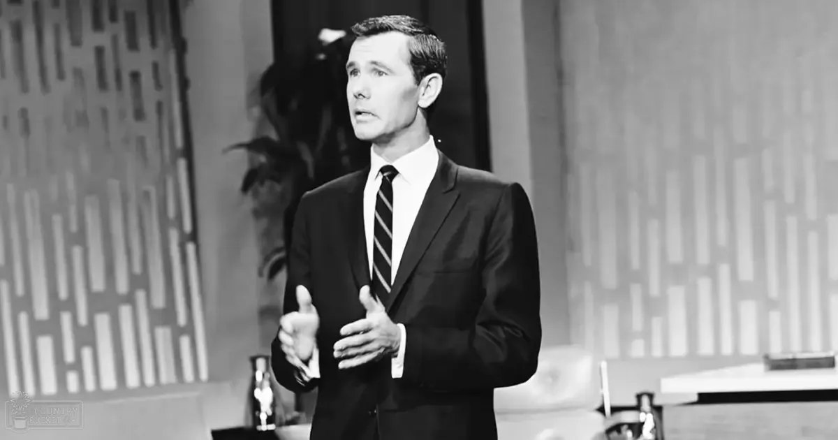 A man in a dark suit standing indoors against a backdrop with geometric patterns and vertical lines; the setting suggests an event or presentation space.