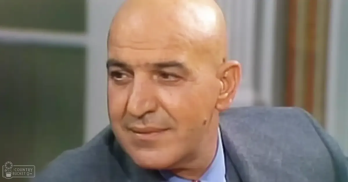 An older adult male wearing a dark gray suit and light blue shirt, standing indoors with neutral-colored curtains and a window frame in the background.