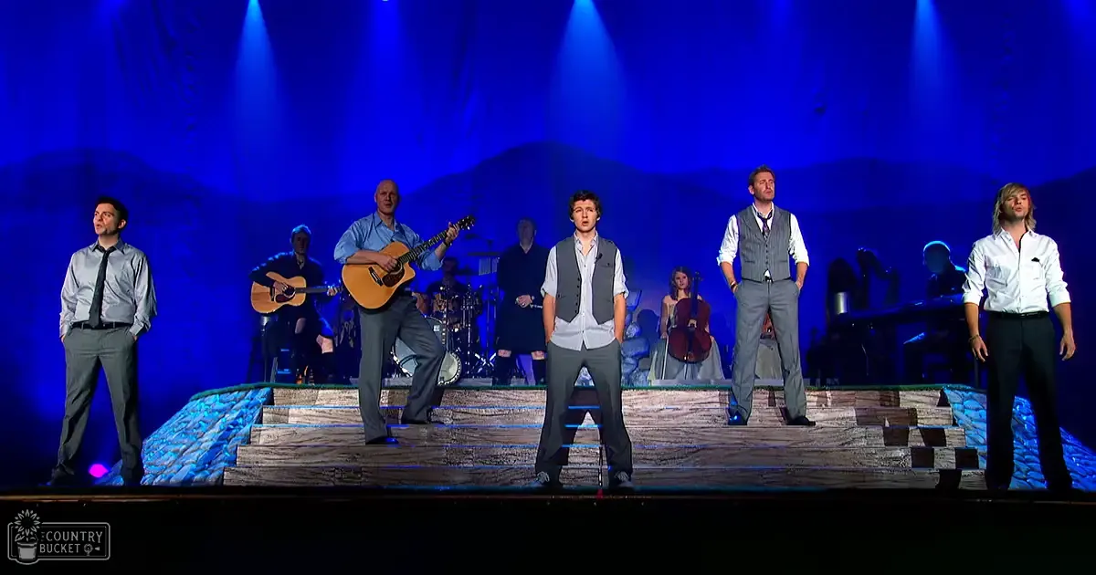 Adult male singer performing in an indoor theater with stage lighting, musical instruments, and a backdrop of mountains under blue lights at night.