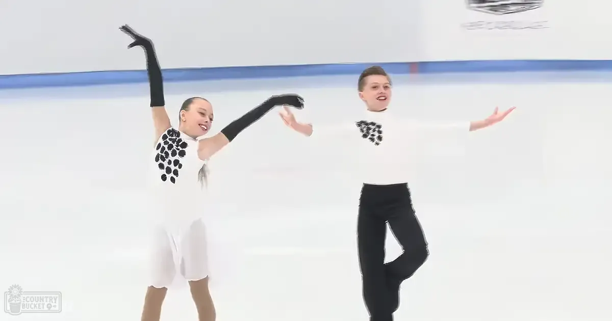 Two young figure skaters performing on an ice rink with white dresses and black polka dots against a backdrop of blue boards.
