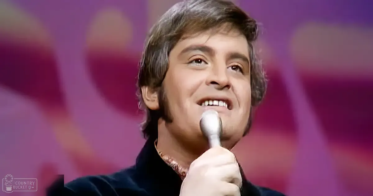 An adult male performer in a dark suit jacket, likely from the mid-20th century, holding a microphone and wearing intricate jewelry against stage lighting with purple and pink hues.