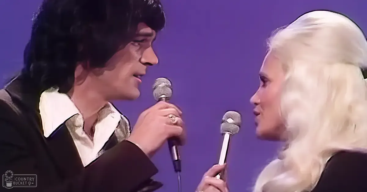 Musical performance scene with two adults, one male in a dark suit jacket with a patterned white shirt and the other female in a shiny white outfit. Setting is an indoor stage with purple lighting.