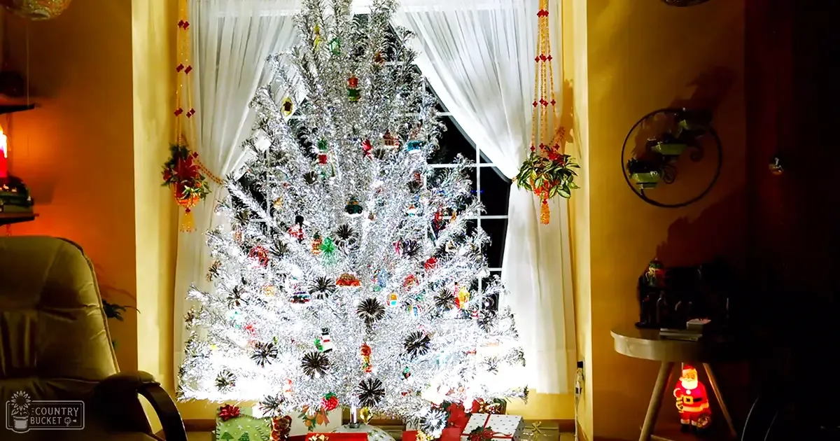 A decorated Christmas tree in a home setting, adorned with silver tinsel and colorful ornaments, surrounded by presents under the tree, framed by curtains and hanging decorations, with a small round table holding a Santa Claus figurine to the right. The scene is indoors, likely during Christmas time.