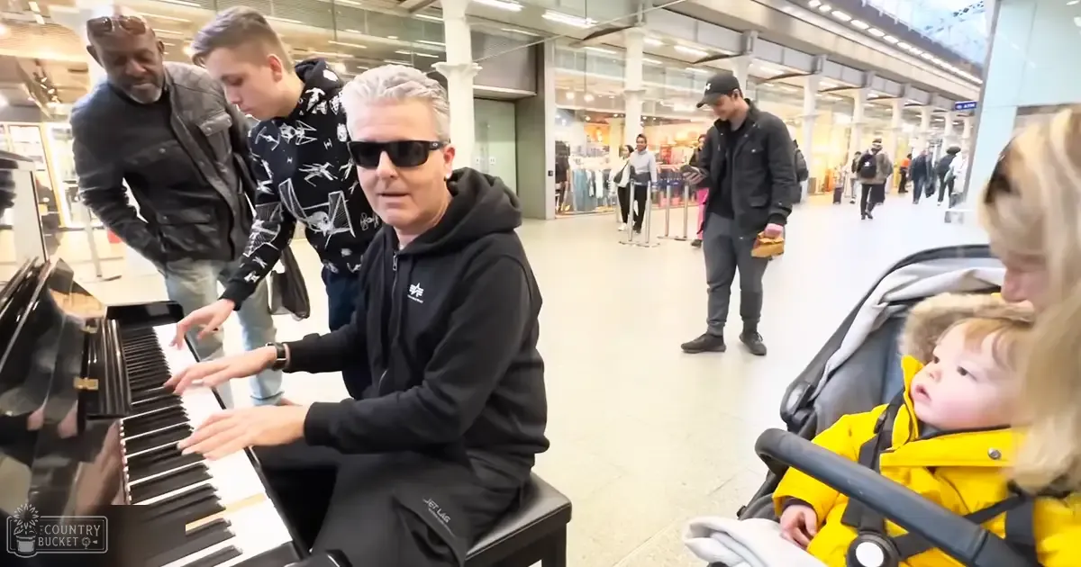 Middle-aged adult male playing piano at an airport terminal, with a young adult male and other travelers in the background under natural light.