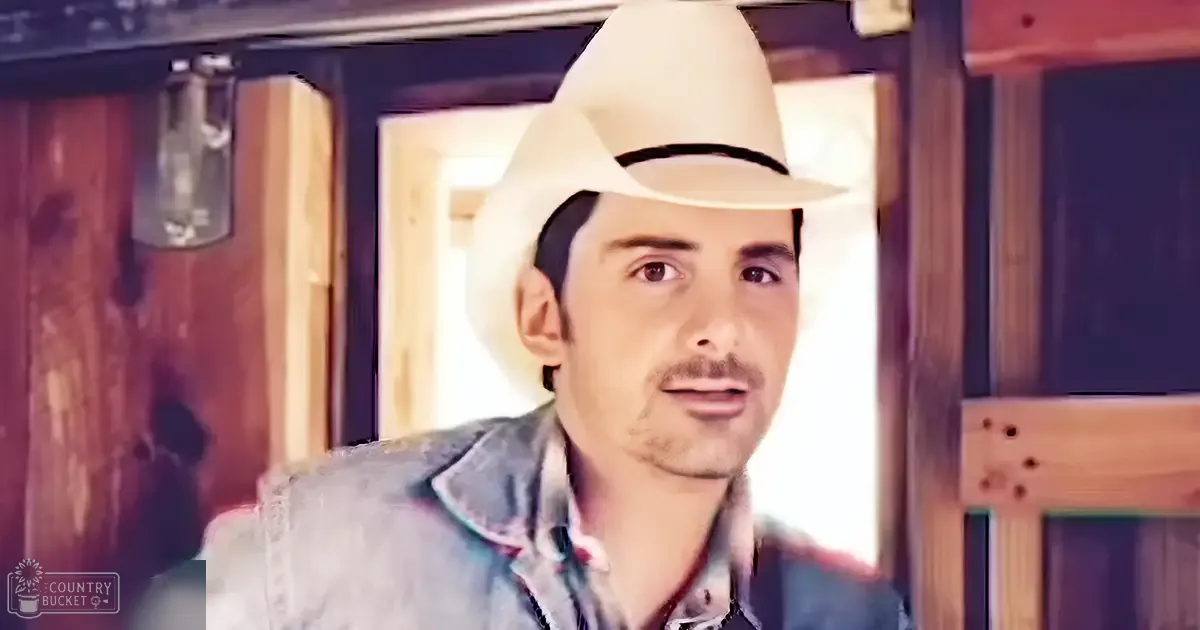A young adult male wearing a light blue denim jacket and patterned shirt stands indoors, surrounded by wooden elements suggesting a rustic cabin setting with natural daylight streaming in.