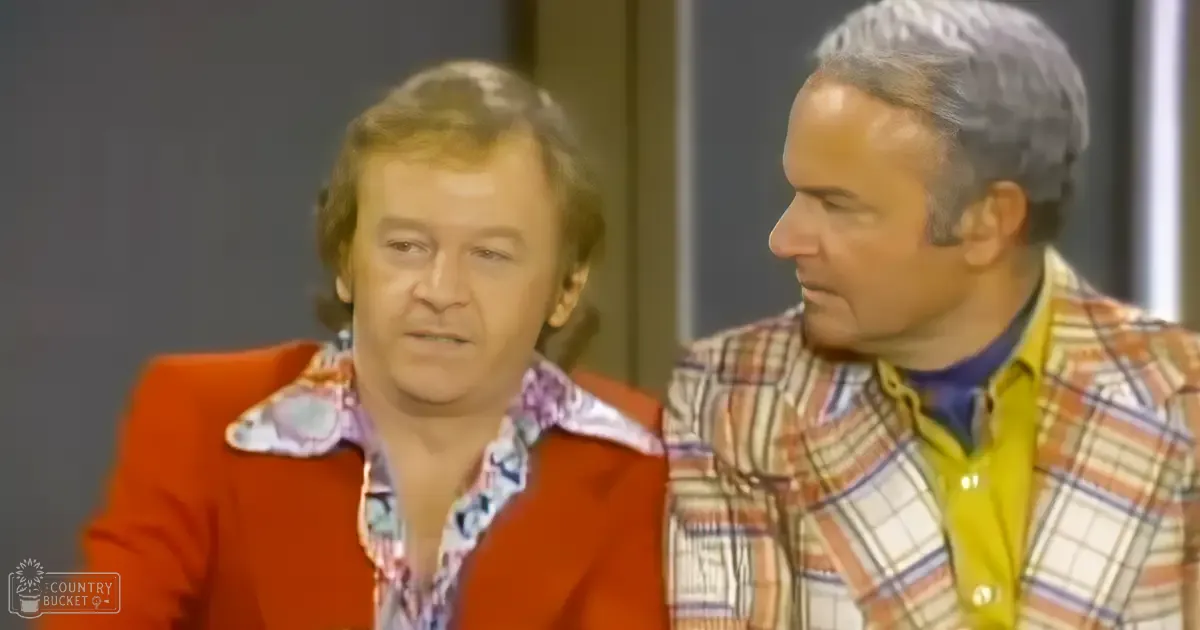 Two middle-aged males wearing vintage-style blazers and shirts in a studio setting with neutral-colored walls, even lighting.